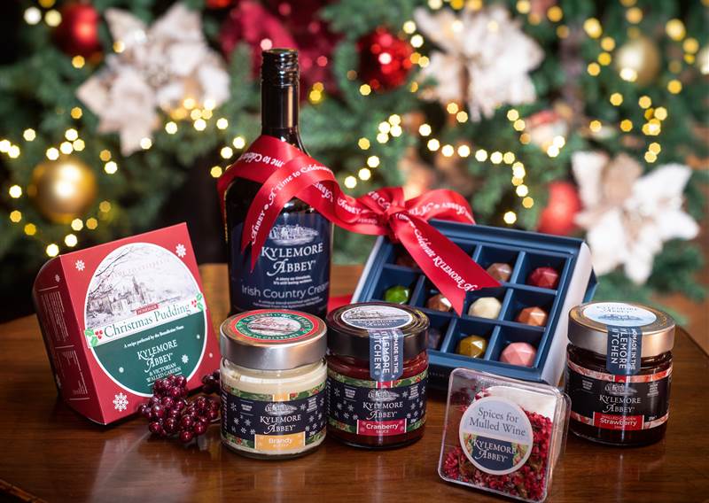 Festive gift set with wine, chocolates, jams, and treats on a table with Christmas lights.